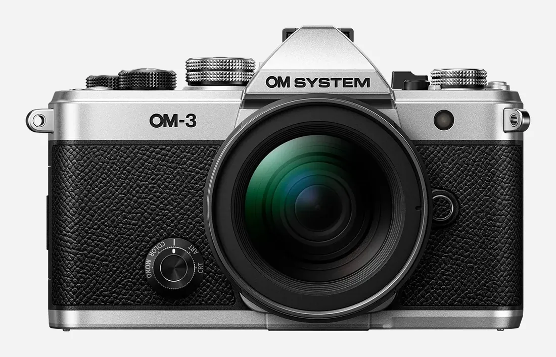 OM SYSTEM OM-3 — OM SYSTEM’in OM-3 sayfasındaki gümüş gövde, bir M.Zuiko zoom objektifle önden görünüm.